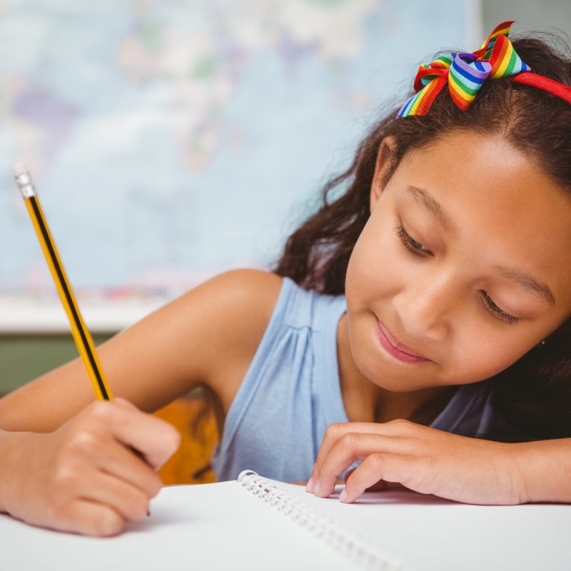 Little girl in a classroom writing in her book with a world map behind her